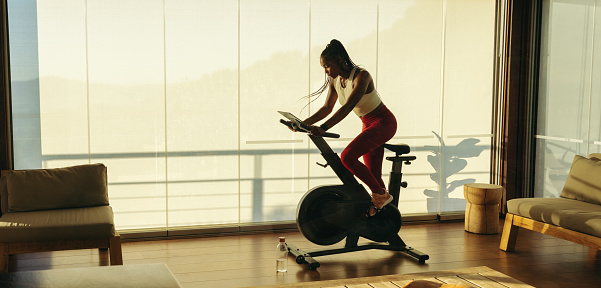 Athletic black woman committing to her fitness routine at home, doing cardio with a smart fitness bike. Young woman working hard to maintain her physical health and achieve her fitness goals.