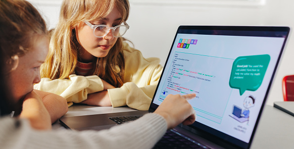 Young female student explaining a computer code to her classmates in a classroom. Group of young children engaging with each other as they learn computer programming skills in a STEM education class.