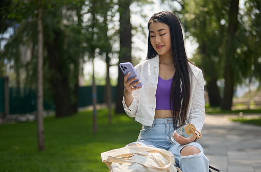 Young South East Asian female person photographed in a daily life with modern smart phone, wireless headphones, trendy glass water bottle and sustainable cotton bag