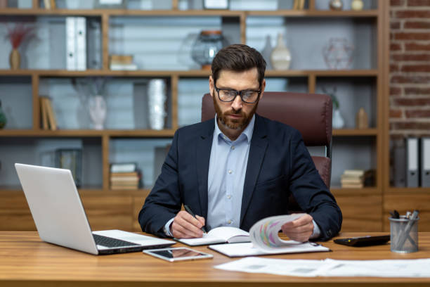 Serious and focused financier accountant on paper work inside office, mature man using calculator and laptop for calculating reports and summarizing accounts, businessman at work