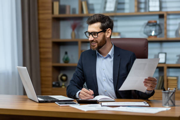 istock Mature businessman in shirt doing paperwork, man working with documents, contracts and bills sitting at table using laptop at work, financier accountant with beard and glasses. 1502450035
