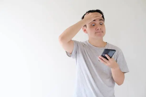 Medium and soft-focused shot of a young Asian man who wears a grey t-shirt and is facepalm while holding while looking at a smartphone. Shallow depth-of-field. Isolated white background.