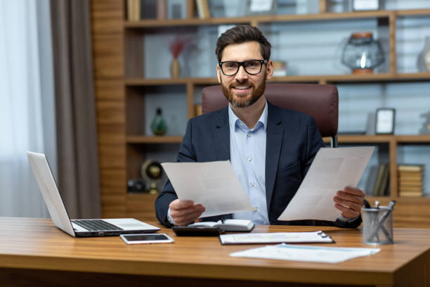istock Portrait of successful financier, man with documents in hands smiling and looking at camera, businessman behind paper work sitting at desk inside office, mature boss in glasses and business suit 1502450370