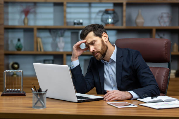 Serious thinking senior businessman working inside office using laptop, man in glasses solving problem, mature investor in business suit