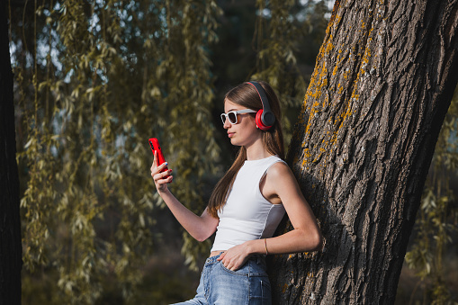 Happy teen with red smartphone and earphones in a park. Happy lifestyle concept. This photograph is perfect for projects related to music, entertainment, youth culture, and personal expression.