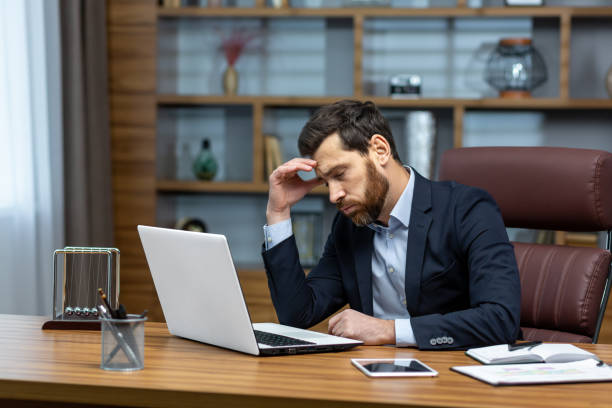 istock sad and depressed businessman in depression working inside modern office building, man in business suit with glasses and beard using laptop for work, boss disappointed with work result. 1502453426
