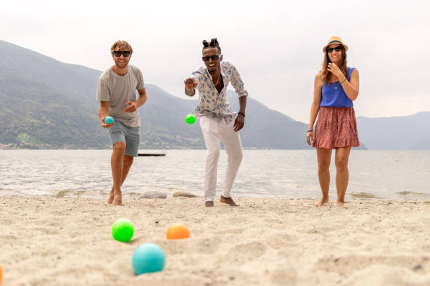 Two petanque sets in black cases with metallic boules and red target balls placed on red concrete edge near sand terrain, evoking relaxed outdoor sport in city environment