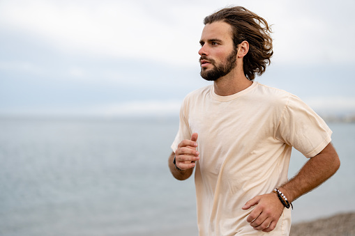 Beautiful young man running on the sandy sea beach in summer wearing a sport clothing, working out outdoors