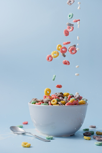 White bowl with colorful ring-shaped cereal and milk. Cereal and milk pouring into the bowl. Milk splashing. Blue background.