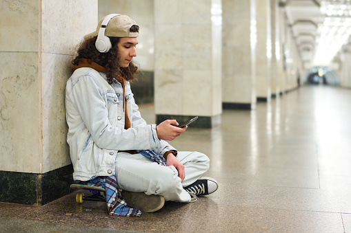 Adolescent guy in casualwear sitting on skateboard by white marble column in subway tunnel and listening to music while waiting for train
