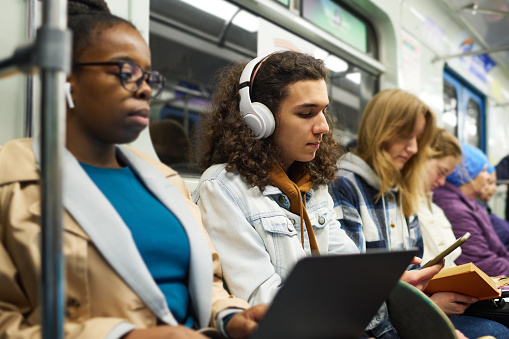 Focus on teenage guy in headphones scrolling through playlist in mobile phone while sitting between two intercultural female passengers