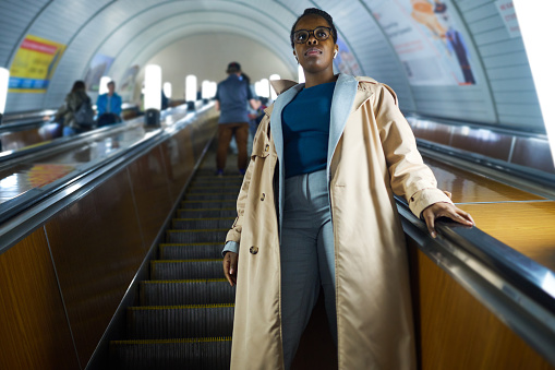 Young African American woman in casualwear standing on escalator and moving downwards against long tunnel of metro with other people