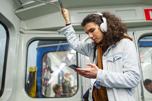 Serious teenage guy in headphones looking at smartphone screen while holding by handrail in train and watching online movie or video