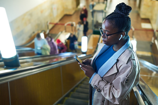 Side view of young black woman in eyeglasses watching online video in smartphone while moving downwards on escalator in metro