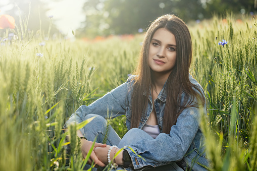 Beautiful brunette girl dressed in jeans is sitting in green cornfield looking at the camera. Horizontally.