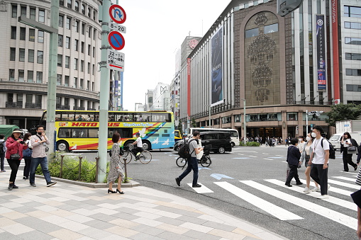 May 26, 2023 - Chuo Ward, Japan: Pedestrians and traffic cross Chuo-dori Avenue, 5 Chome, in the upscale Ginza shopping district. The famous Mitsukoshi department store stands to the right. Spring morning with hazy skies.