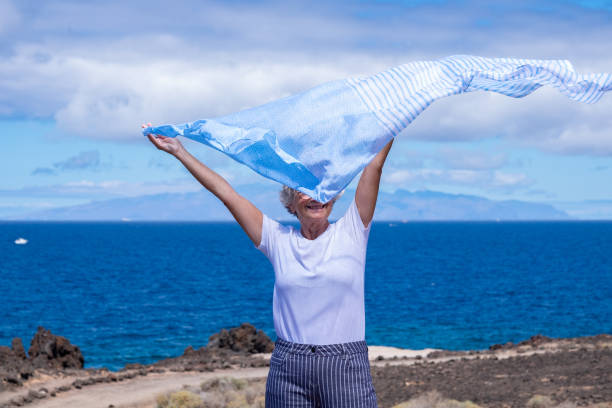 femme âgée heureuse et active en vacances en mer tout en agitant une écharpe dans le ciel avec les bras tendus. fond bleu - senior adult outdoors wellbeing sky photos et images de collection