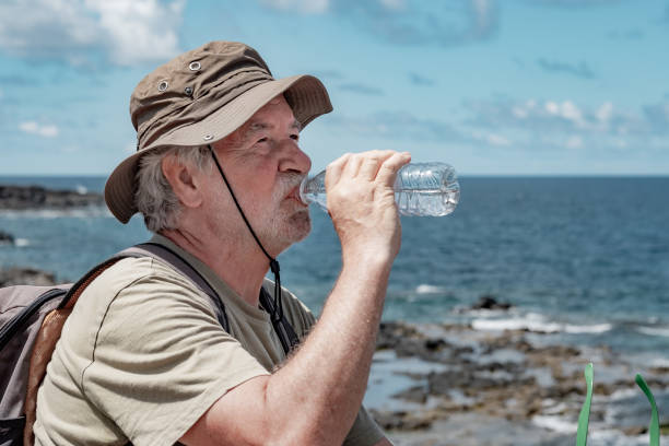 istock Senior bearded man with hat and backpack sitting on the rock beach drinking a water bottle, elderly grandfather looking away enjoying freedom and holidays 1502488382