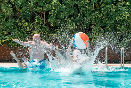 Four senior people in sport activity jumping into the swimming pool playing with inflatable ball. Elderly couples women and men enjoying holidays or retirement