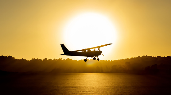 Backlit silhouette of a cessna plane outlined with the sun just behind it flying low with the sea water below illuminated by the golden light of sunset and trees and vegetation in the background