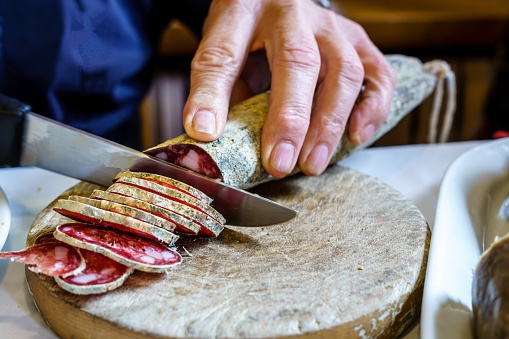 Hands of a man cutting fuet with a knife on wood. fuet, espetec, tastet, petador or secallona is a typical sausage of the Catalan gastronomy, Spain.