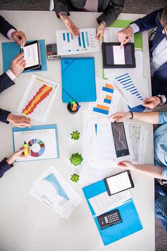 Bussy table at a meeting with digital tablet, calculators, graphs, documents lying around. Man looking at a chart on his tablet.