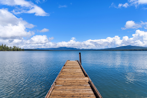 A long wooden boat ramp extending onto a blue mountain lake under Spring blue sky and white clouds. Crane Prairie Reservoir, Bend, Oregon, USA.