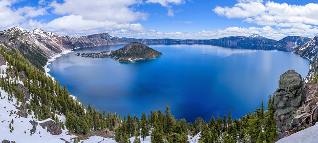 A panoramic cliff-side overview of deep blue Crater Lake, surrounded by the rugged lake rim, on a calm Spring day. Crater Lake National Park, Oregon, USA.