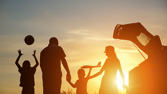Family silhouettes spend time together in countryside near open car boot. Siblings holding parents hands play with toys at back sunset on blue sky