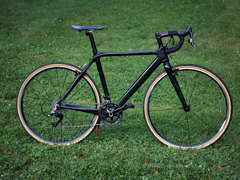 black carbon bicycle in field of grass (road bike with drop handlebars, skinwall tires) cyclocross gravel bike sleek, new build (700c wheels, double crankset, cantilever brakes, shifters)