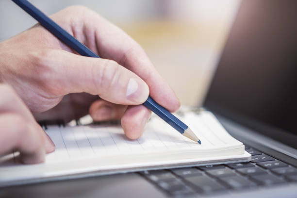 istock Focused view of a man's hand as he writes in a notepad on a modern laptop, background gently blurred 1502537669