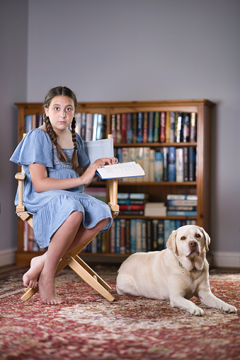Eleven-year-old girl wearing a blue dress reading in her library with her pet dog by her side.