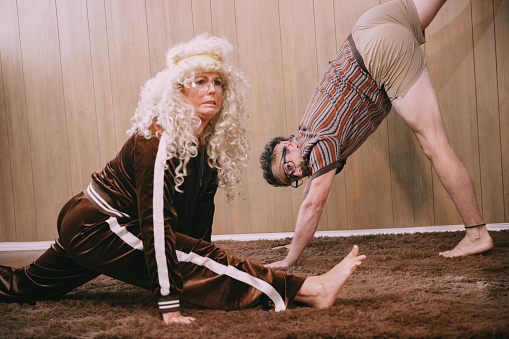 A middle aged couple in late 1970's - early 1980's vintage clothing, exercise and do stretches in front of a wood veneer paneling on a brown shag carpet / rug.   Humor concept with fun and funny faces.  Brown color tones.
