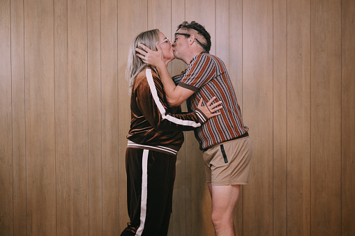 A middle aged couple in late 1970's - early 1980's vintage clothing embrace and kiss in front of a wood veneer paneling on a brown shag carpet / rug.   Humor concept with fun and funny faces.  Brown color tones.