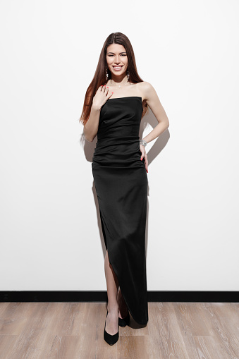 Full body of confident smiling young female model in elegant black dress standing against white background and looking at camera with wearing high heels shoes