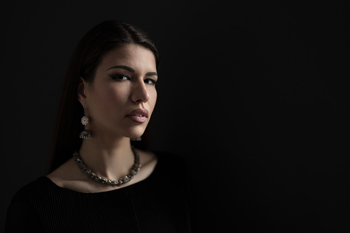 Portrait of attractive young female model wearing black dress with necklace and earrings looking at camera against dark background in darkness