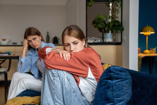 Frustrated teenage girl sits hugging his knees, looks away. Mom sits next daughter and comforts after quarrel. Communication problems between parent and kids.Ignoring children problems.Bulling school
