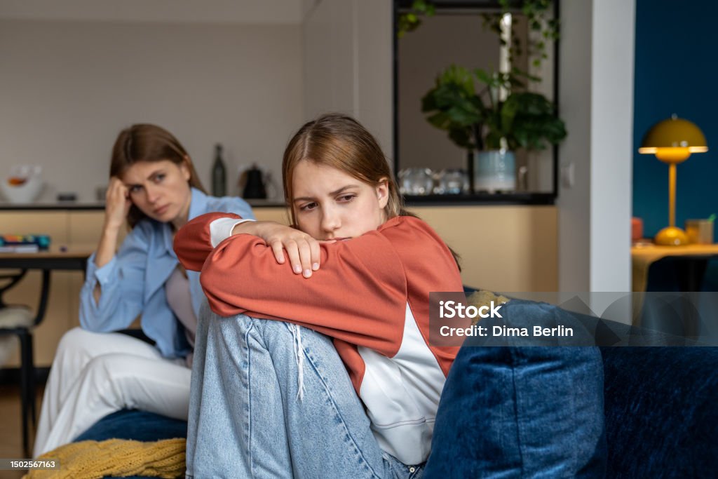 Frustrated offended teenage girl sits hugging knees and looks away. Mom sits next daugther Frustrated teenage girl sits hugging his knees, looks away. Mom sits next daughter and comforts after quarrel. Communication problems between parent and kids.Ignoring children problems.Bulling school Mother Stock Photo