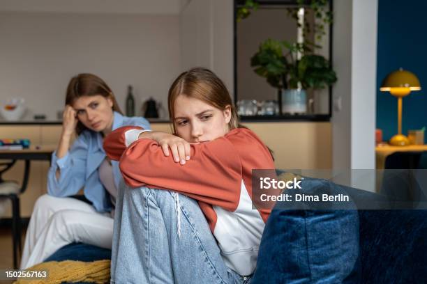 Frustrated Offended Teenage Girl Sits Hugging Knees And Looks Away Mom Sits Next Daugther Stock Photo - Download Image Now