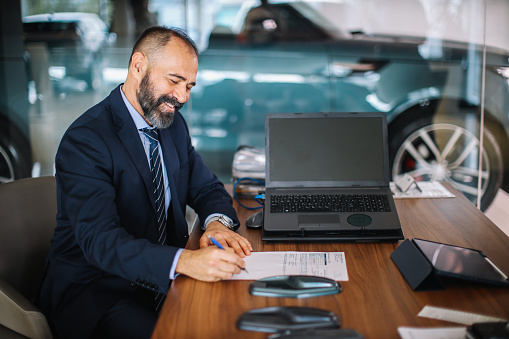 Mid age businessman visits the car dealership, looking at the current offers, talking to the salesperson. He is sitting at the desk, choosing which colour they like the best, signing the paperwork, buying a new car.