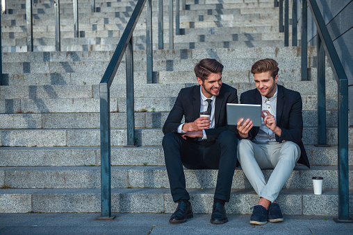 Two handsome young businessmen in formal clothes are using a tablet and smiling while sitting on stairs of the office building during coffee break
