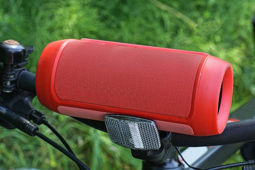 one red mobile plastic music speaker lies on the black metal handlebar of a sports bike on the street against the background of green vegetation