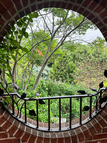 Looking through the round window on this brick building, you have a great scenic view of a beautiful garden with lovely green trees and plants.