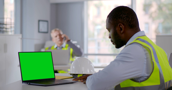 Side view of African-American engineer in reflective vest looking at laptop screen in office. Green screen. Foreman having video conference with contractor