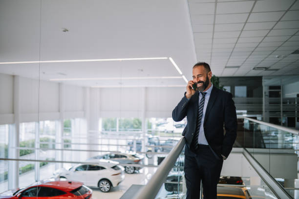 mid aged man working in the car showroom Mid aged man working in the luxury car showroom - dealership. He is checking his mobile phone, answering calls and emails, going through everyday stuff. bmw dealership stock pictures, royalty-free photos & images