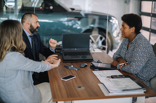 Mid aged couple visits the car dealership, looking at the current offers, talking to the salesperson. They’re sitting at the desk, choosing which colour they like the best, finishing paperwork, buying a new car.