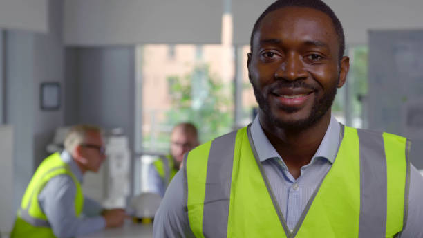 istock African foreman in reflective vest looking at camera standing in modern office. Portrait of positive architect looking at camera with colleagues working on background 1502582671