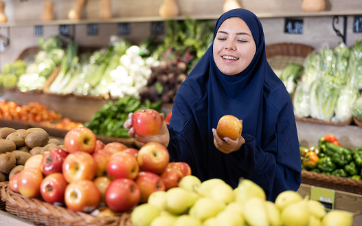 Girl in paranja making purchases in supermarket, choosing fresh ripe apples