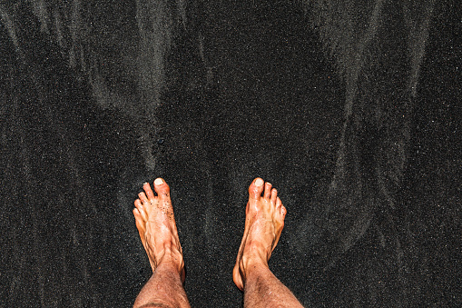 High contrast image of mans feet standing on black sand beach in Maui in Honokalani, Maui, Hawaii.