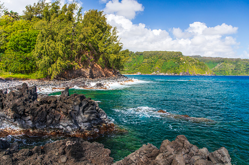 Coastal scene of rocks and blue ocean with green mountains on the road to Hana in Maui, Hawaii.